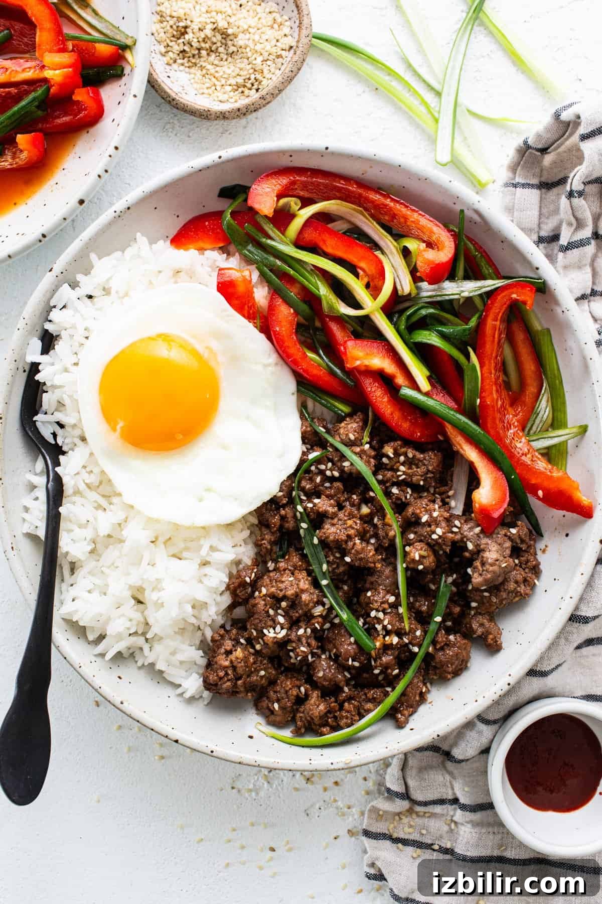 A plate of rice topped with a fried egg, stir-fried beef, and sliced bell peppers, accompanied by condiments on a white surface.