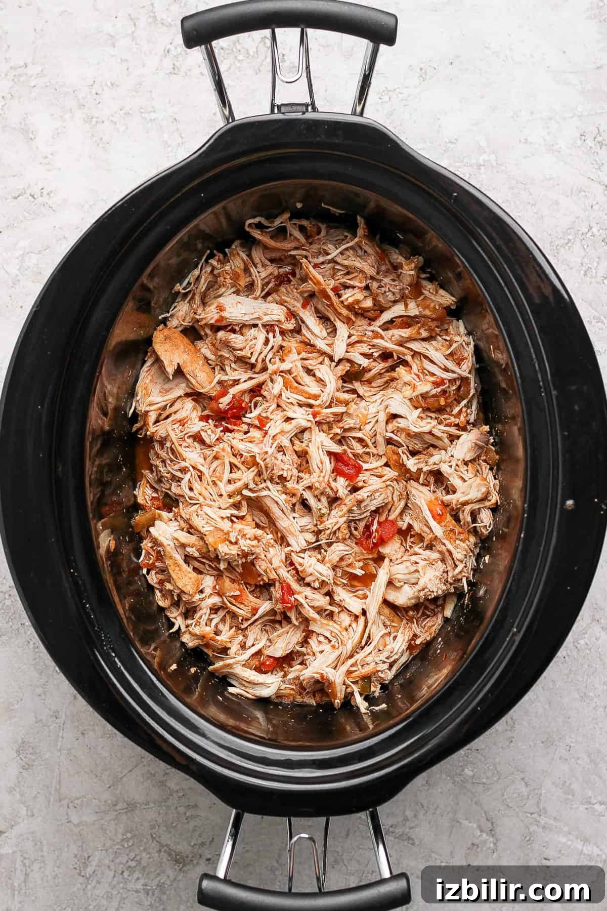 Shredded chicken mixed with red peppers in a slow cooker on a gray textured surface.