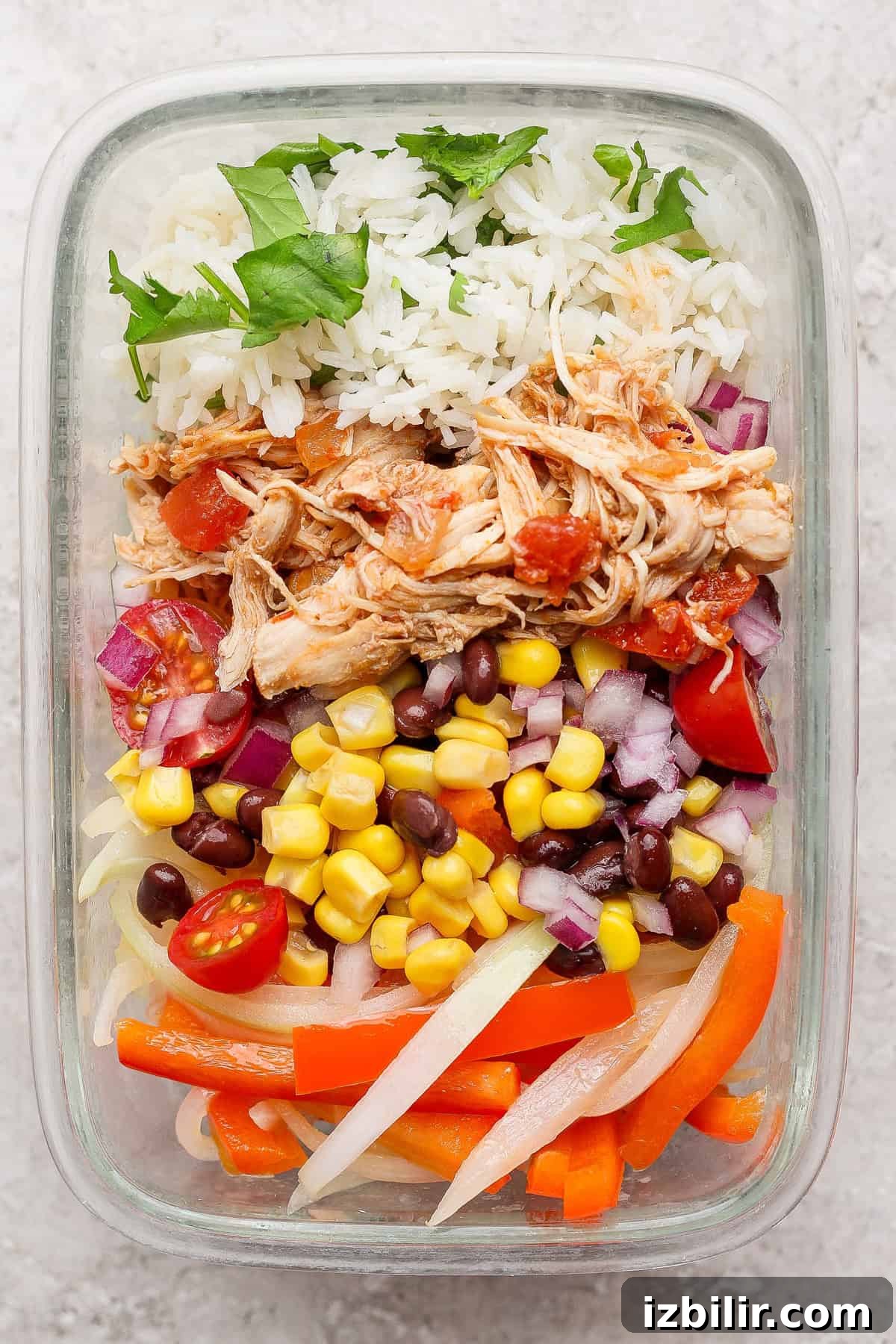 A glass container with rice, shredded chicken, black beans, corn, red onions, cherry tomatoes, sliced bell peppers, and cilantro.