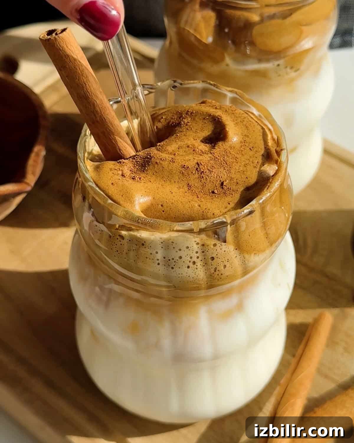 Pumpkin Spice Dalgona Coffee Recipe 6 A glass of whipped coffee with a thick foam layer on top, garnished with a cinnamon stick, sits on a wooden board next to a second similar drink.