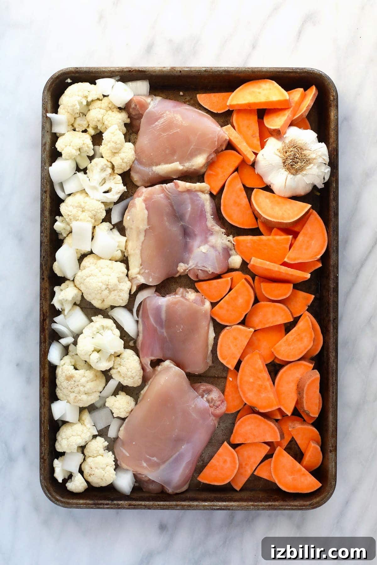 Raw chicken, cauliflower, sweet potatoes, onion, and garlic on a baking sheet ready to bake.