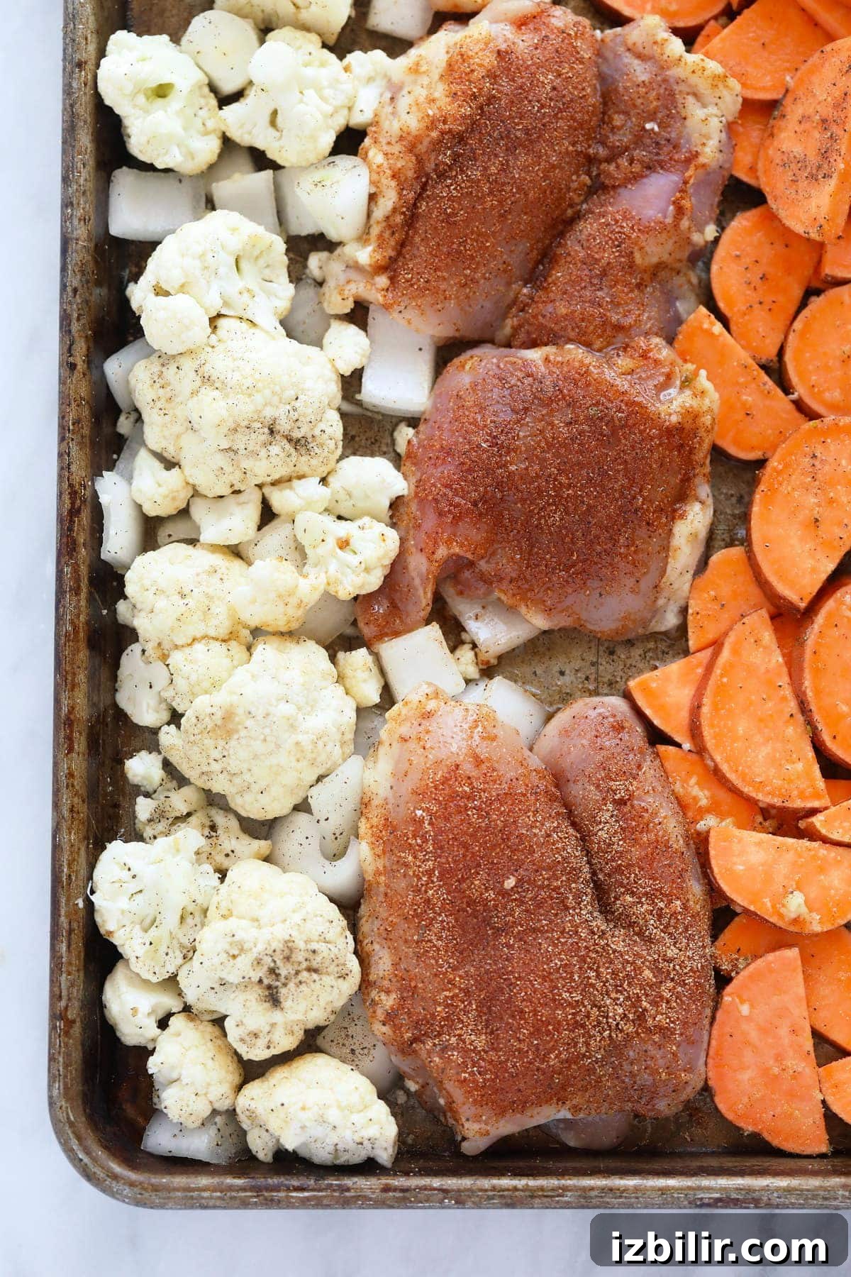 Chicken thighs, sweet potatoes, cauliflower, and onion on a baking sheet.