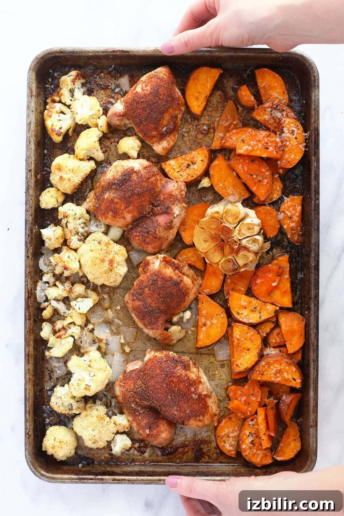 Chicken thighs, sweet potatoes, cauliflower, and garlic on a baking sheet.
