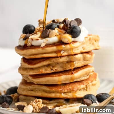 A stack of pancakes topped with blueberries, granola, yogurt, chocolate chips, and syrup being poured over them.