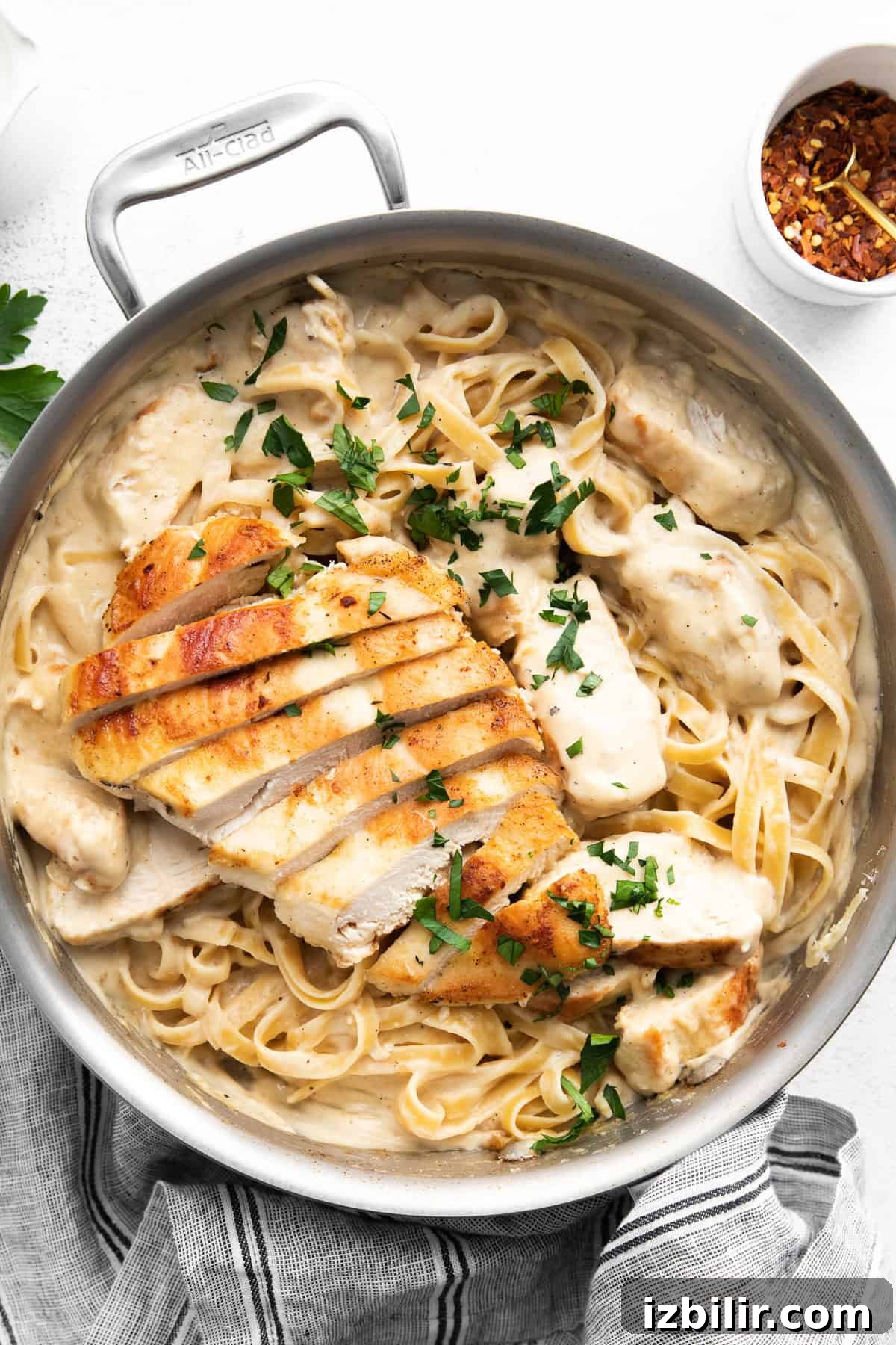 Homemade Creamy Chicken Alfredo 2 A pan filled with creamy fettuccine pasta topped with slices of grilled chicken and garnished with parsley. A small bowl of red pepper flakes is seen in the background.