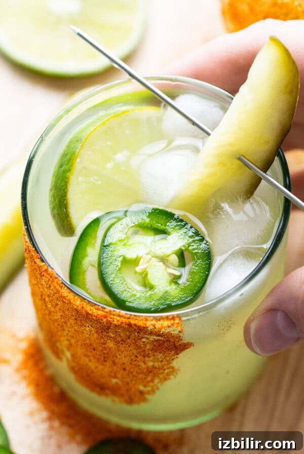 A hand holding a glass of drink with a pickle and lime.