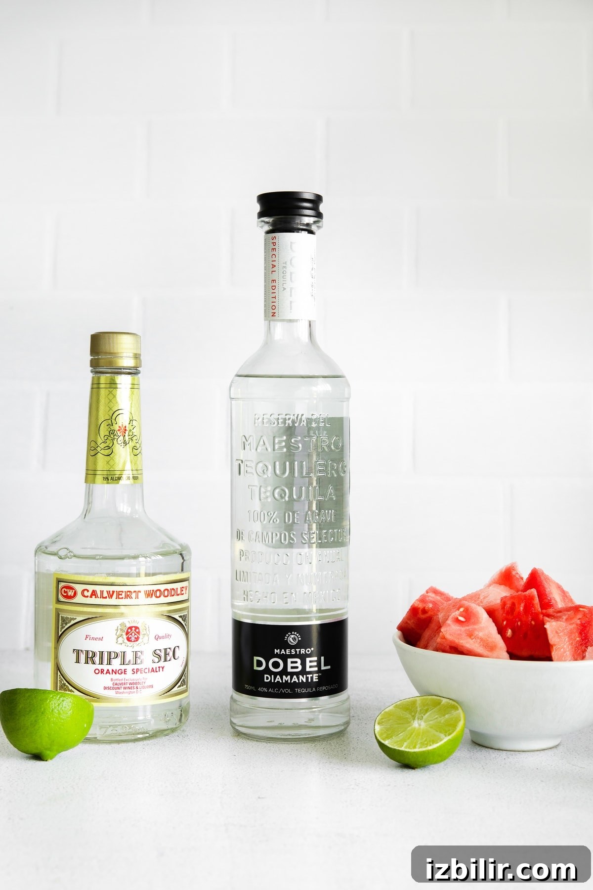 Triple Sec, tequila, fresh watermelon and lime wedges on a counter top.