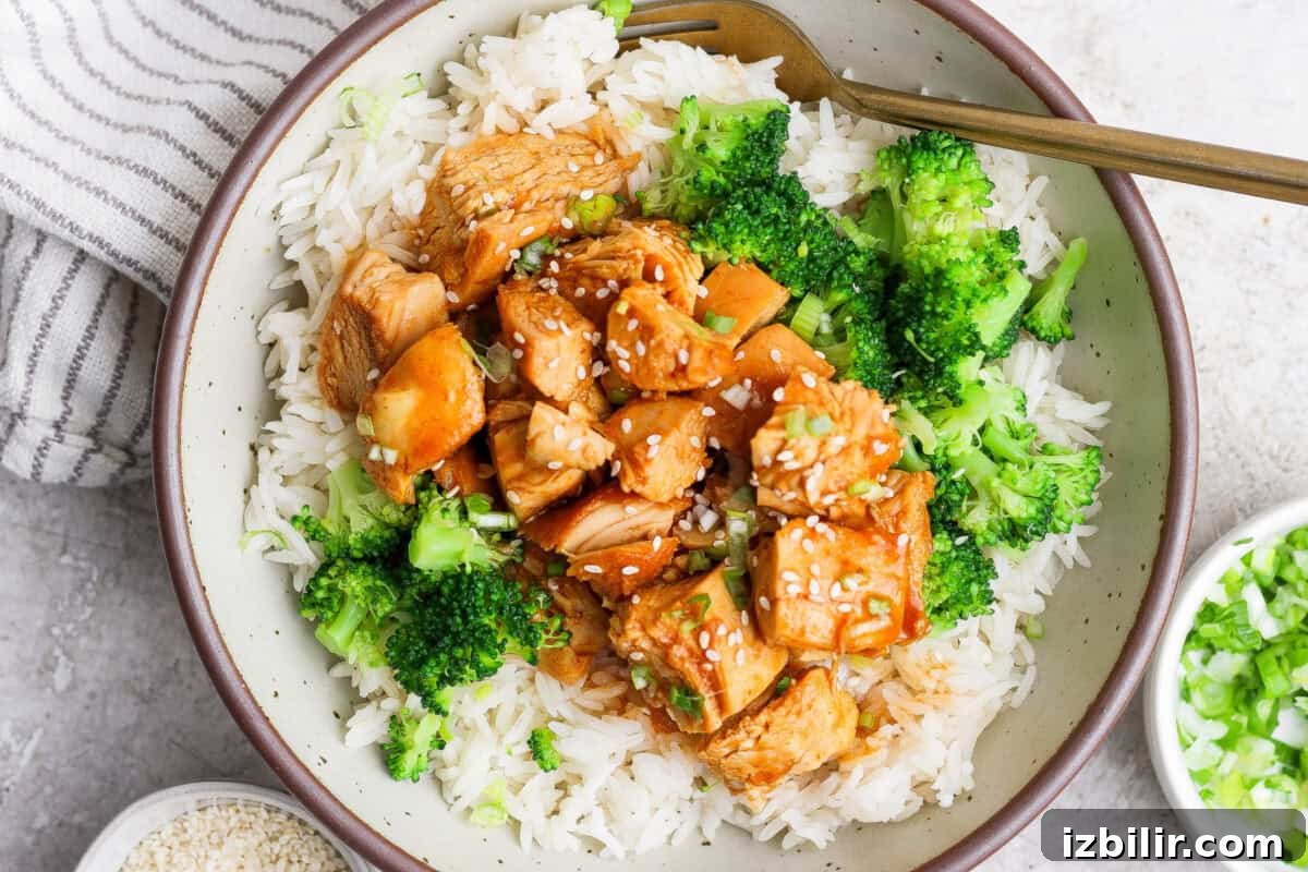 A bowl of white rice topped with broccoli, diced chicken in a brown sauce, and sprinkled with sesame seeds, with a fork on the side.