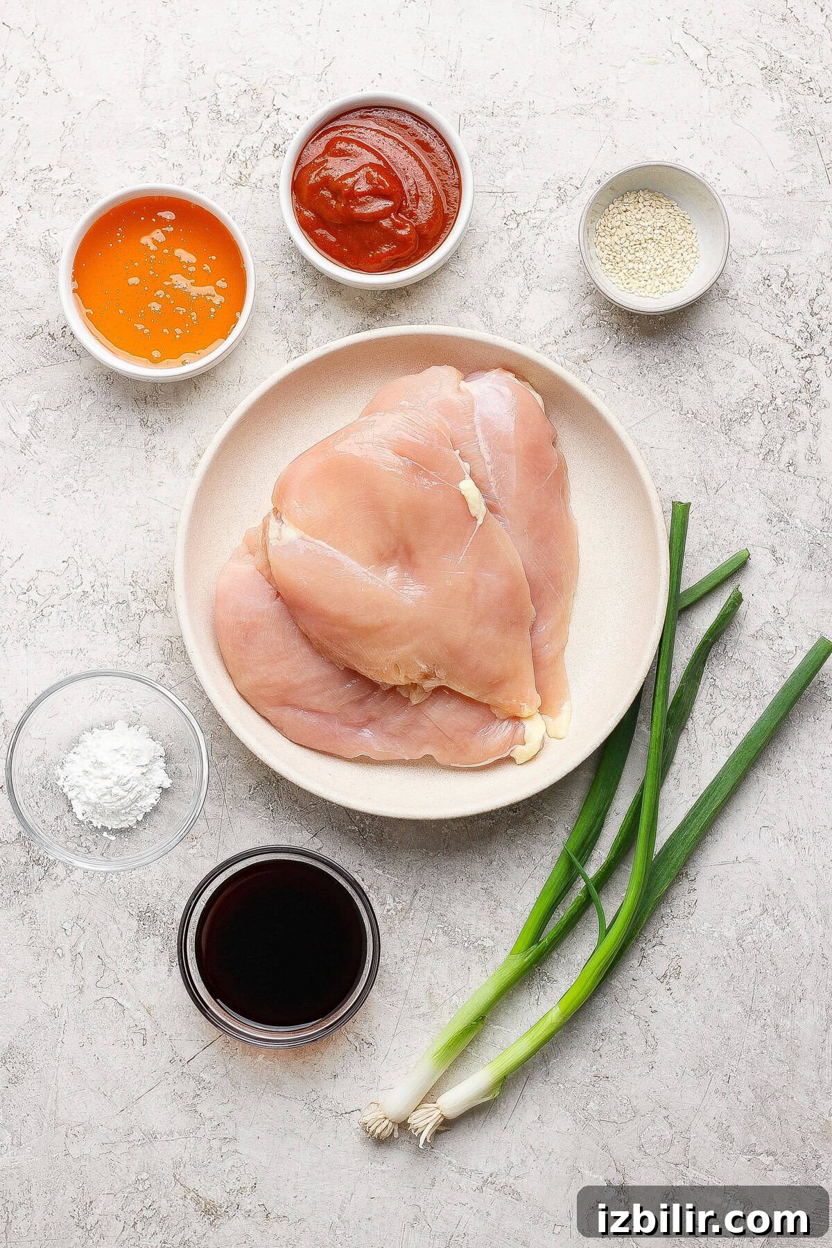 Raw chicken breasts on a plate surrounded by bowls of orange sauce, red sauce, sesame seeds, cornstarch, soy sauce, and two green onions on a light surface.