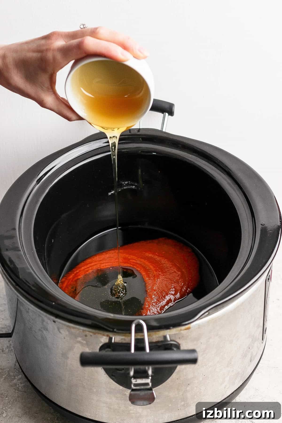 A hand pours a light brown liquid from a cup into a slow cooker containing a red sauce.