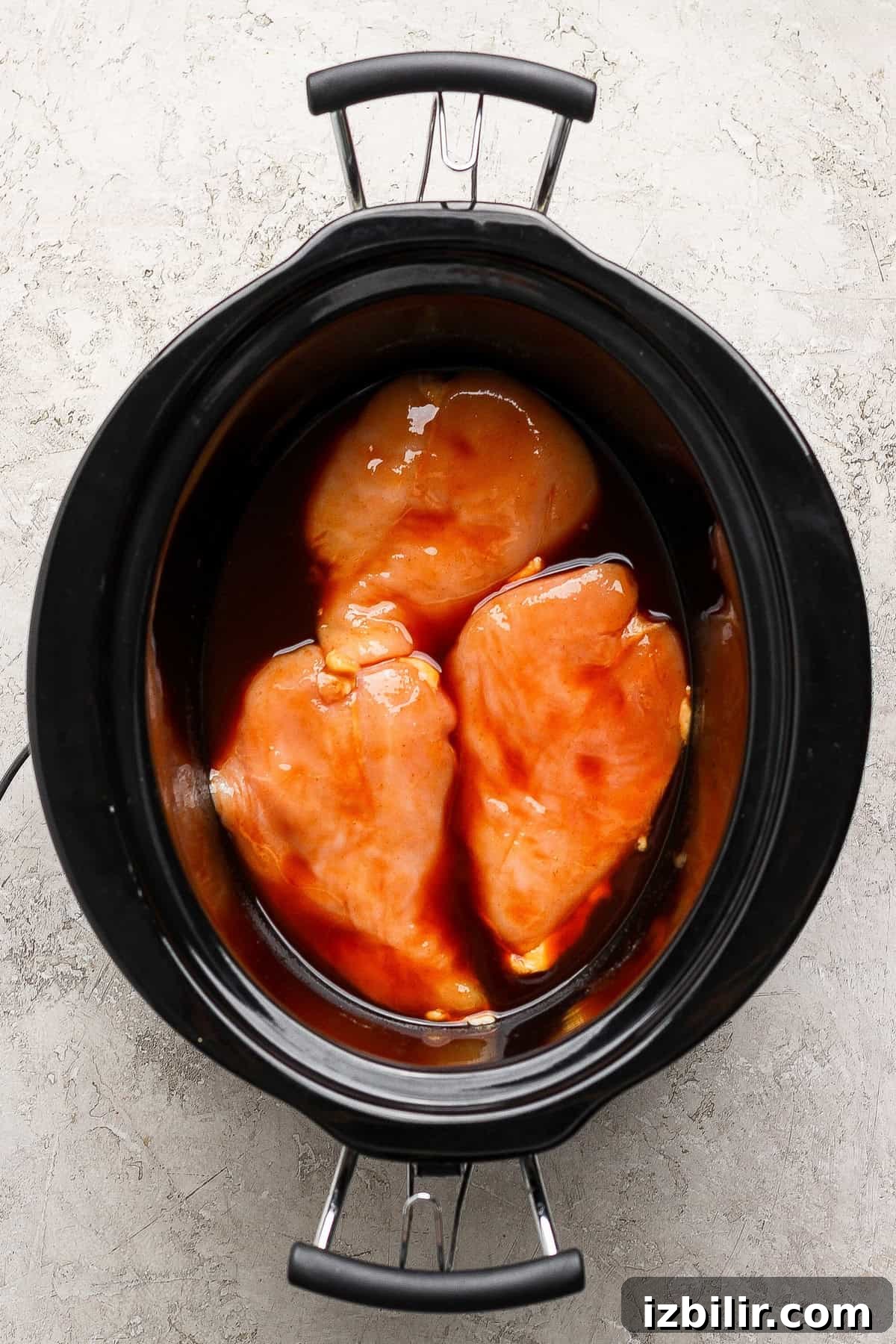 Three raw chicken breasts coated in sauce are placed in a black slow cooker on a light textured surface.