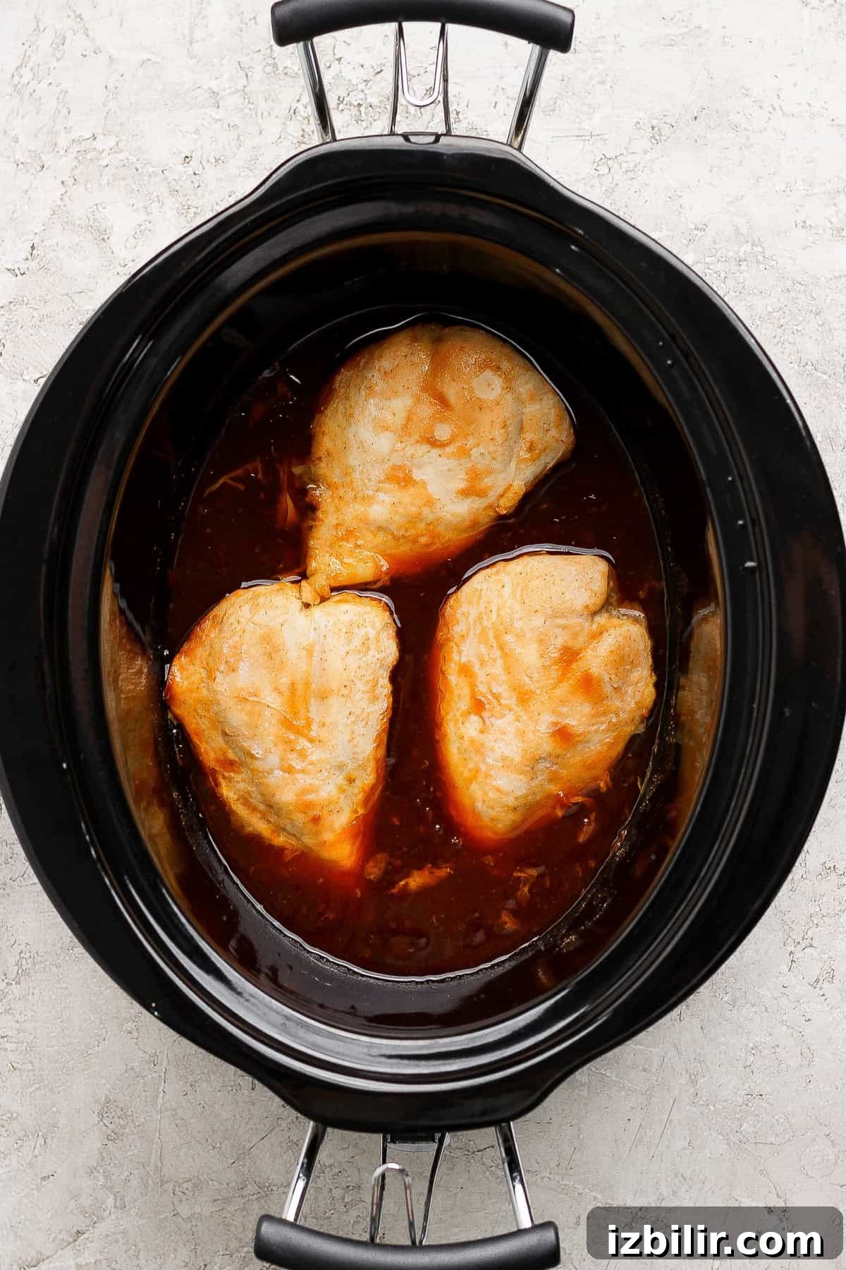 Three pieces of chicken breast cooking in a slow cooker with a dark brown sauce.