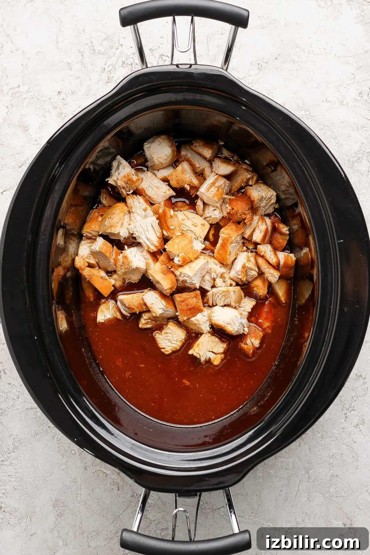 Diced cooked chicken in a slow cooker with a reddish-brown sauce, viewed from above on a light-colored surface.