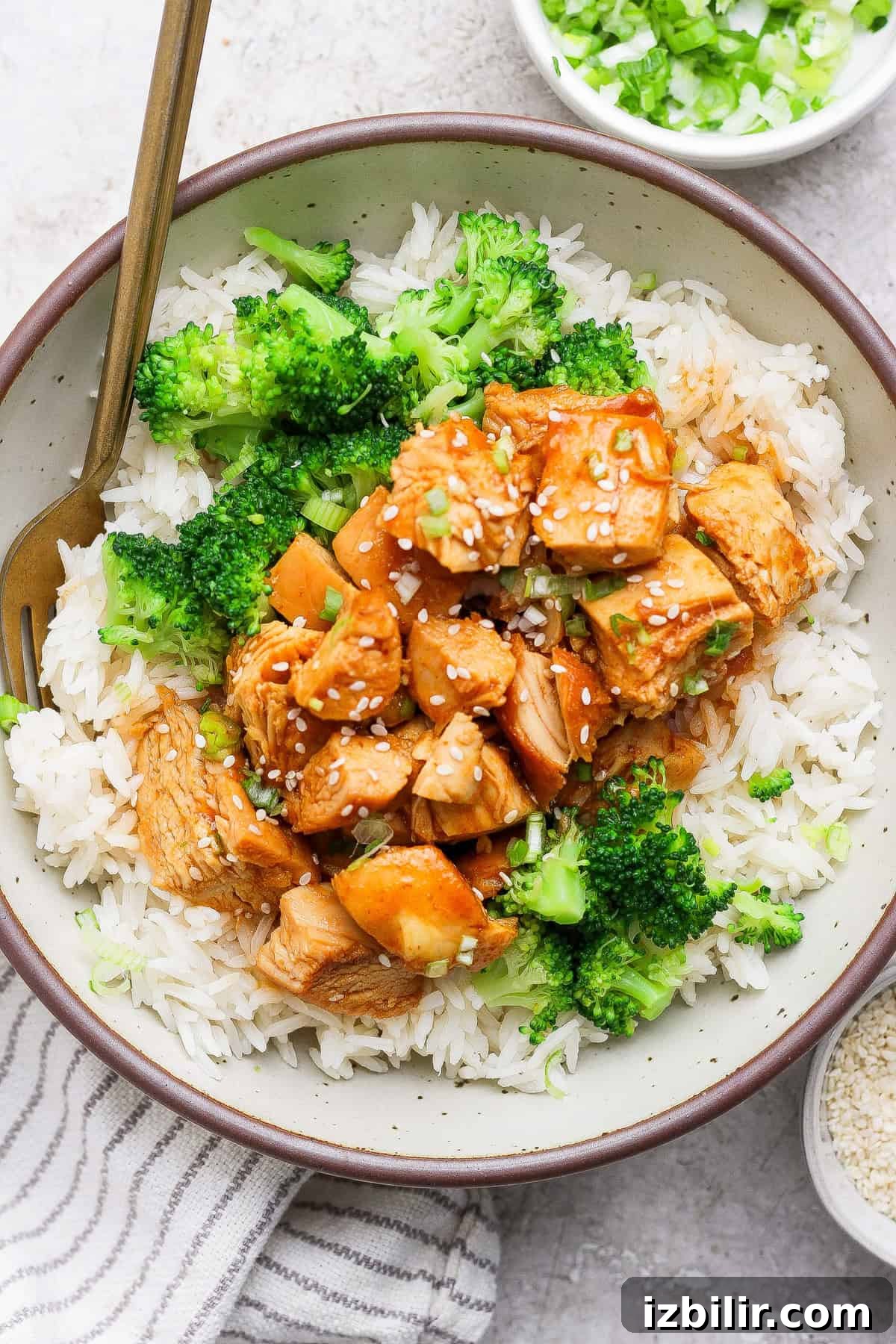 A bowl of white rice topped with broccoli and chunks of sauced chicken, garnished with sesame seeds, with a fork resting in the bowl.