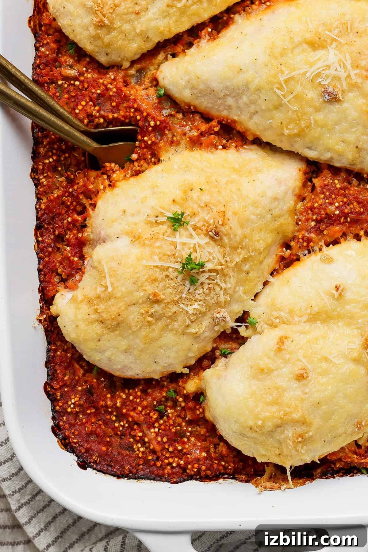 Baked chicken breasts topped with cheese on a bed of tomato quinoa in a white baking dish, garnished with herbs and cheese.