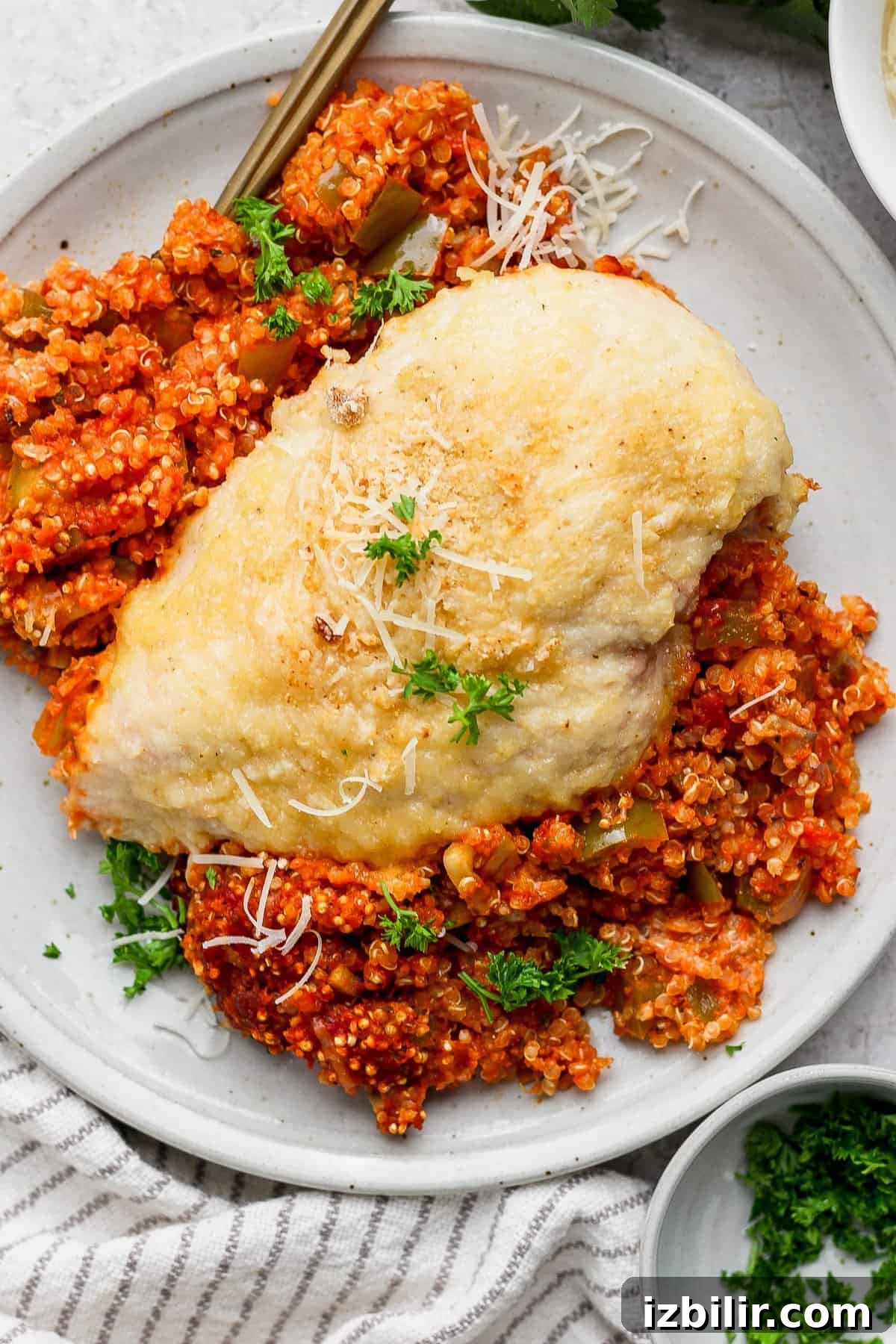 Grilled chicken breast on a bed of tomato quinoa, garnished with grated cheese and parsley on a white plate.