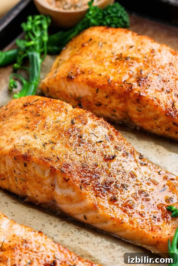 Three golden broiled salmon fillets on a parchment-lined baking sheet with broccolini and lemon.