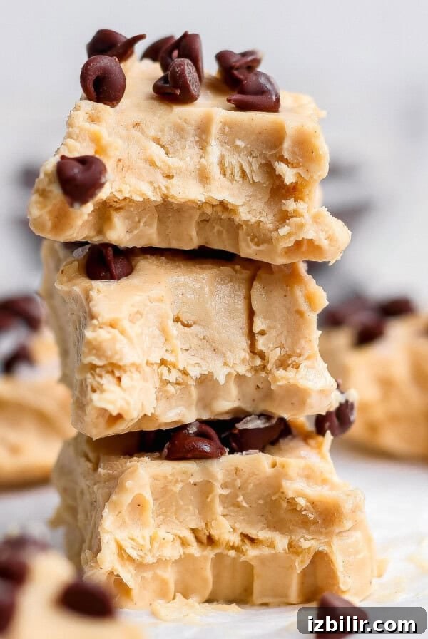Three pieces of peanut butter fudge stacked on top of each other, each topped with chocolate chips, with more fudge pieces in the background.