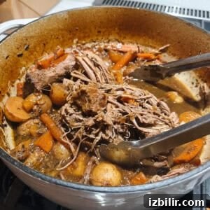 Oven Braised Classic Pot Roast with Red Wine and Root Vegetables 2 A pot of savory pot roast with tender shredded beef, carrots, and potatoes in flavorful broth. Tongs rest on the edge of the pot.