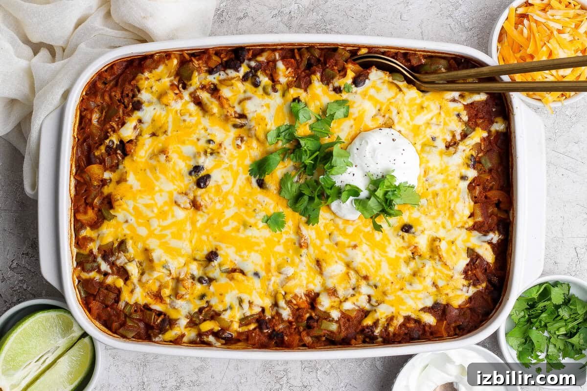 One-Pan Chicken Enchilada Casserole Recipe 2 A baked casserole topped with melted cheese, black beans, sour cream, and chopped cilantro, surrounded by bowls of lime, shredded cheese, and extra sour cream.