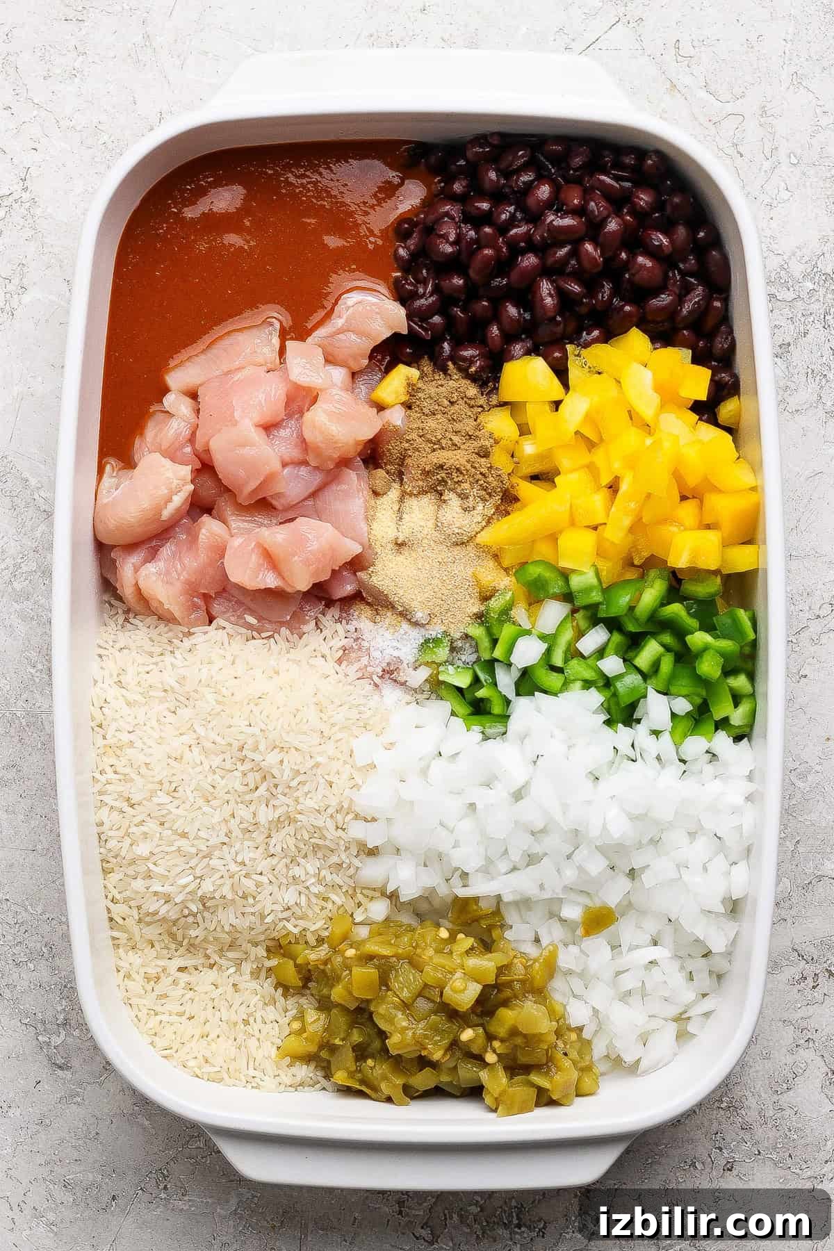 One-Pan Chicken Enchilada Casserole Recipe 3 A casserole dish with raw chicken, black beans, enchilada sauce, yellow bell pepper, green bell pepper, chopped onions, green chilies, rice, and seasonings arranged in sections.
