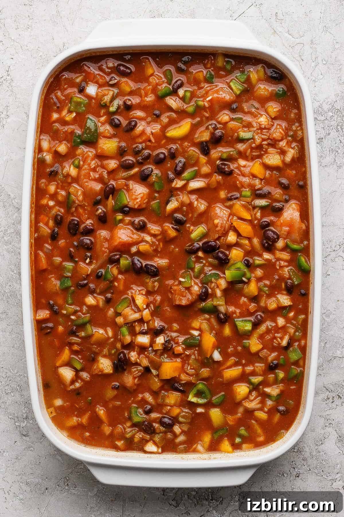 One-Pan Chicken Enchilada Casserole Recipe 4 A rectangular white casserole dish filled with a chunky mixture of beans, tomatoes, rice, and vegetables in a red sauce on a textured surface.