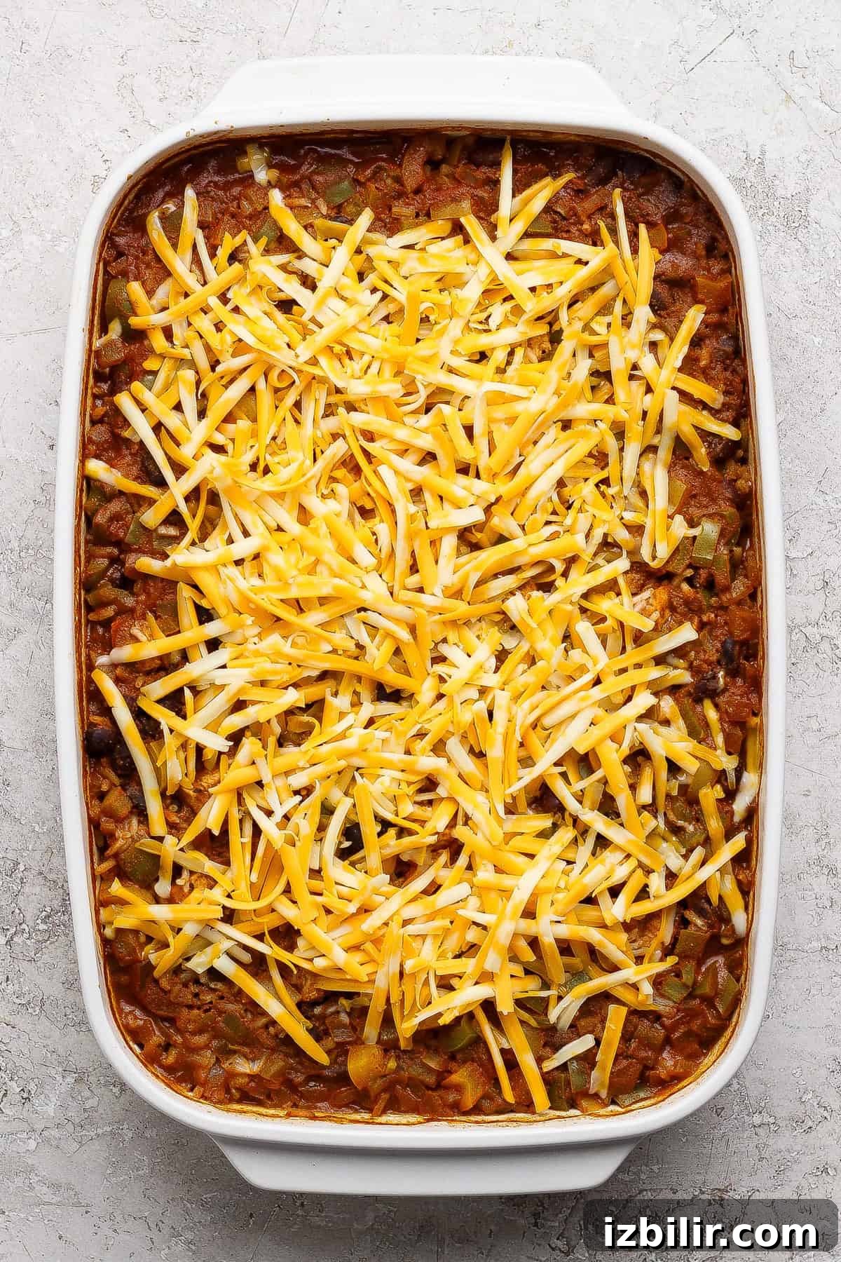 One-Pan Chicken Enchilada Casserole Recipe 5 A rectangular baking dish filled with a casserole topped with a layer of shredded yellow and white cheese, set on a light textured surface.
