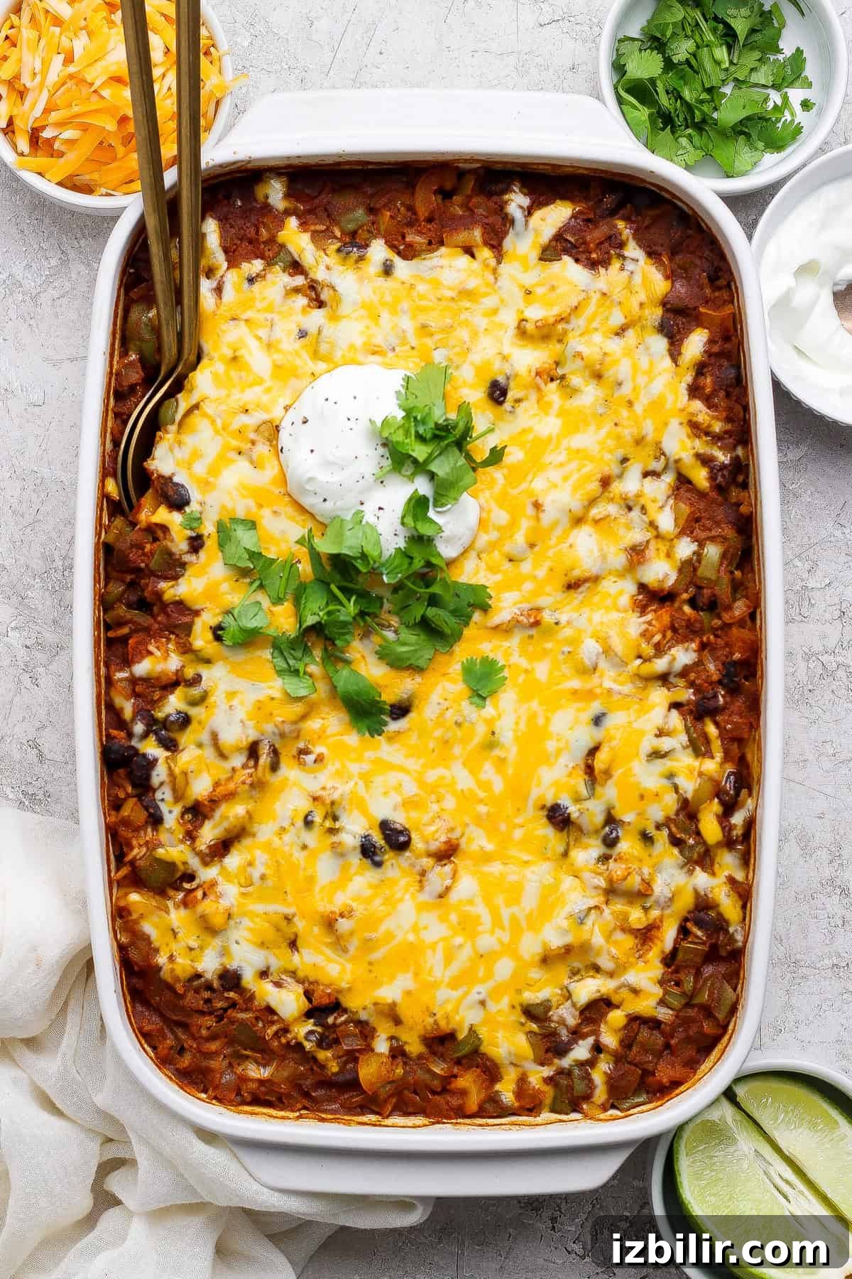 One-Pan Chicken Enchilada Casserole Recipe 6 A baked casserole topped with melted cheese, sour cream, and fresh cilantro, in a white dish, with lime wedges, shredded cheese, and sour cream in bowls on the side.