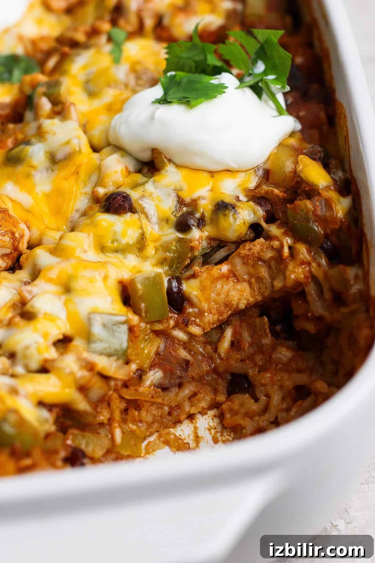 One-Pan Chicken Enchilada Casserole Recipe 7 A baked casserole with melted cheese, rice, beans, and green peppers, topped with a dollop of sour cream and fresh cilantro in a white dish.