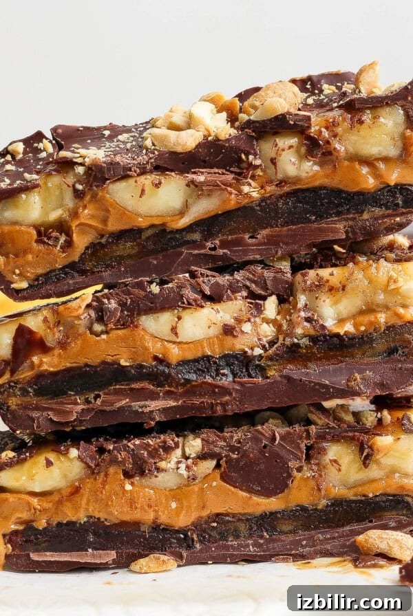 Three stacked pieces of chocolate bark filled with layers of peanut butter, banana slices, and chopped nuts on a parchment paper surface.