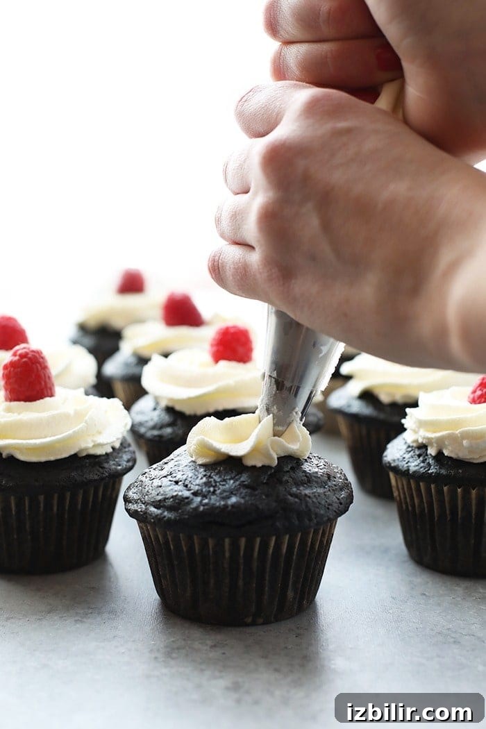 Healthier chocolate cupcakes being piped with vanilla frosting and topped with a fresh raspberry