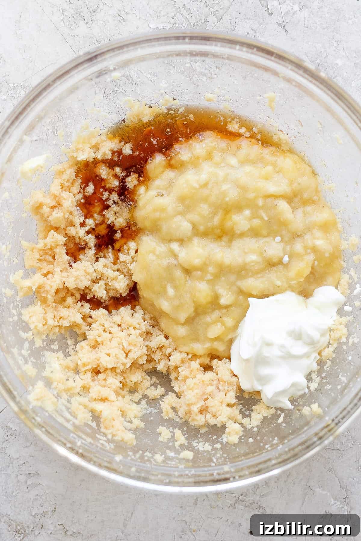Ingredients combined in a glass bowl, possibly for baking, including mashed bananas, sugars, and a dollop of sour cream or yogurt.