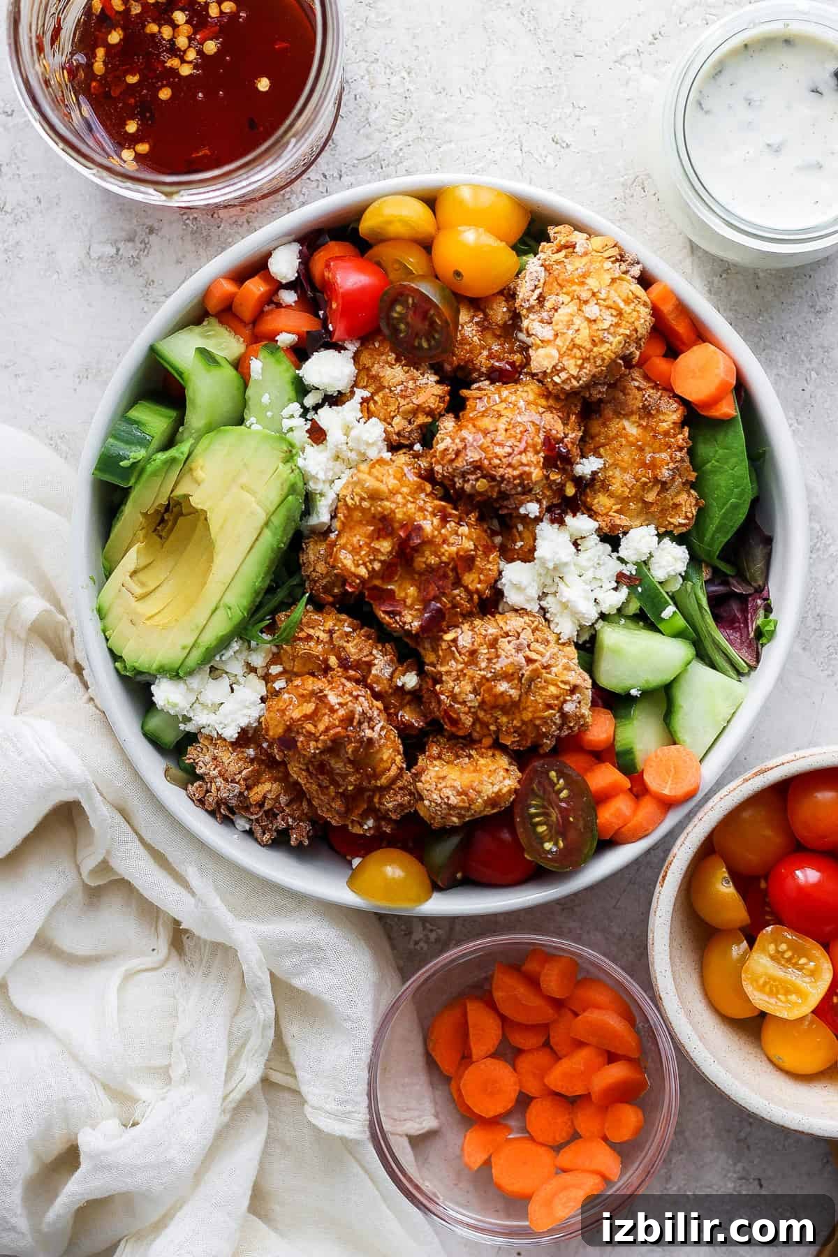 A colorful salad bowl with quinoa, mixed greens, avocado slices, cherry tomatoes, cucumbers, carrots, and topped with crumbled cheese and breaded chicken pieces, accompanied by two small bowls.