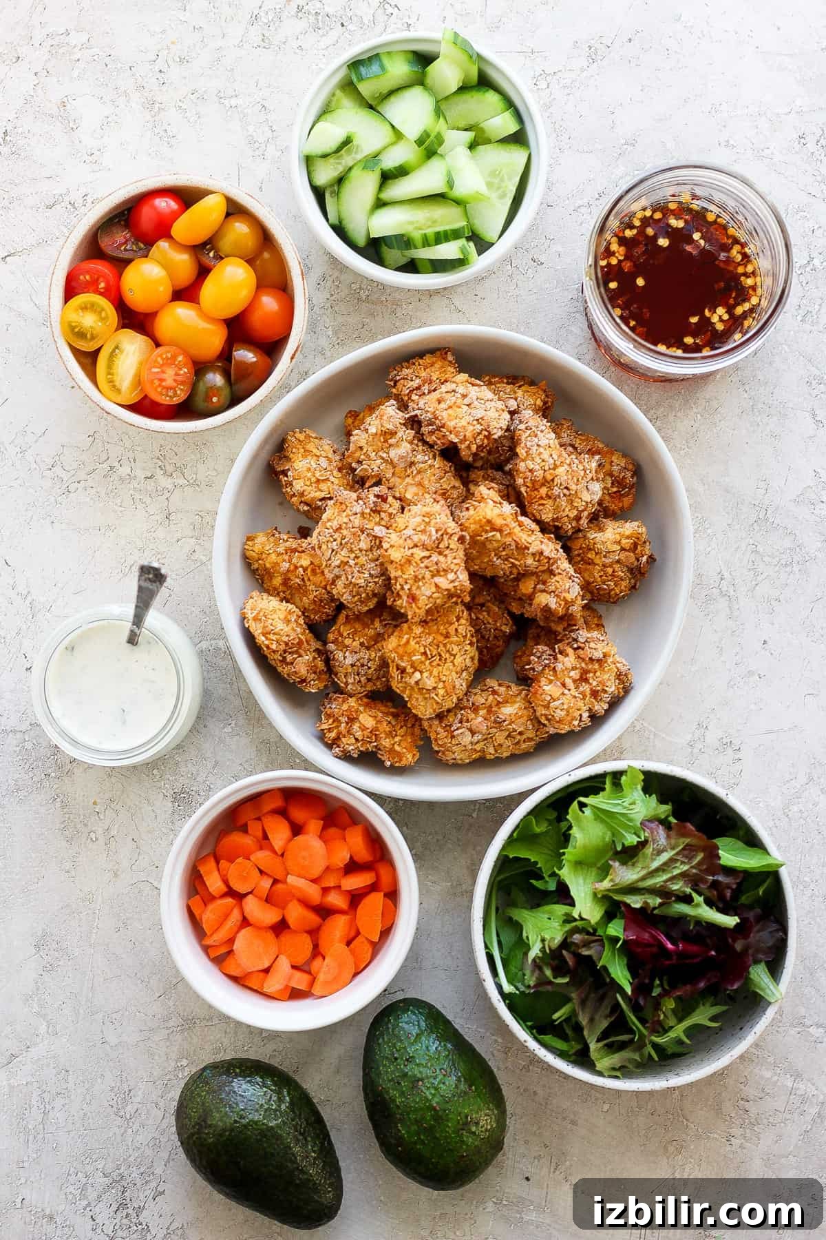 Assorted healthy food ingredients with breaded chicken pieces, bowls of fresh vegetables, mixed greens, avocado, and dips arranged on a light surface.