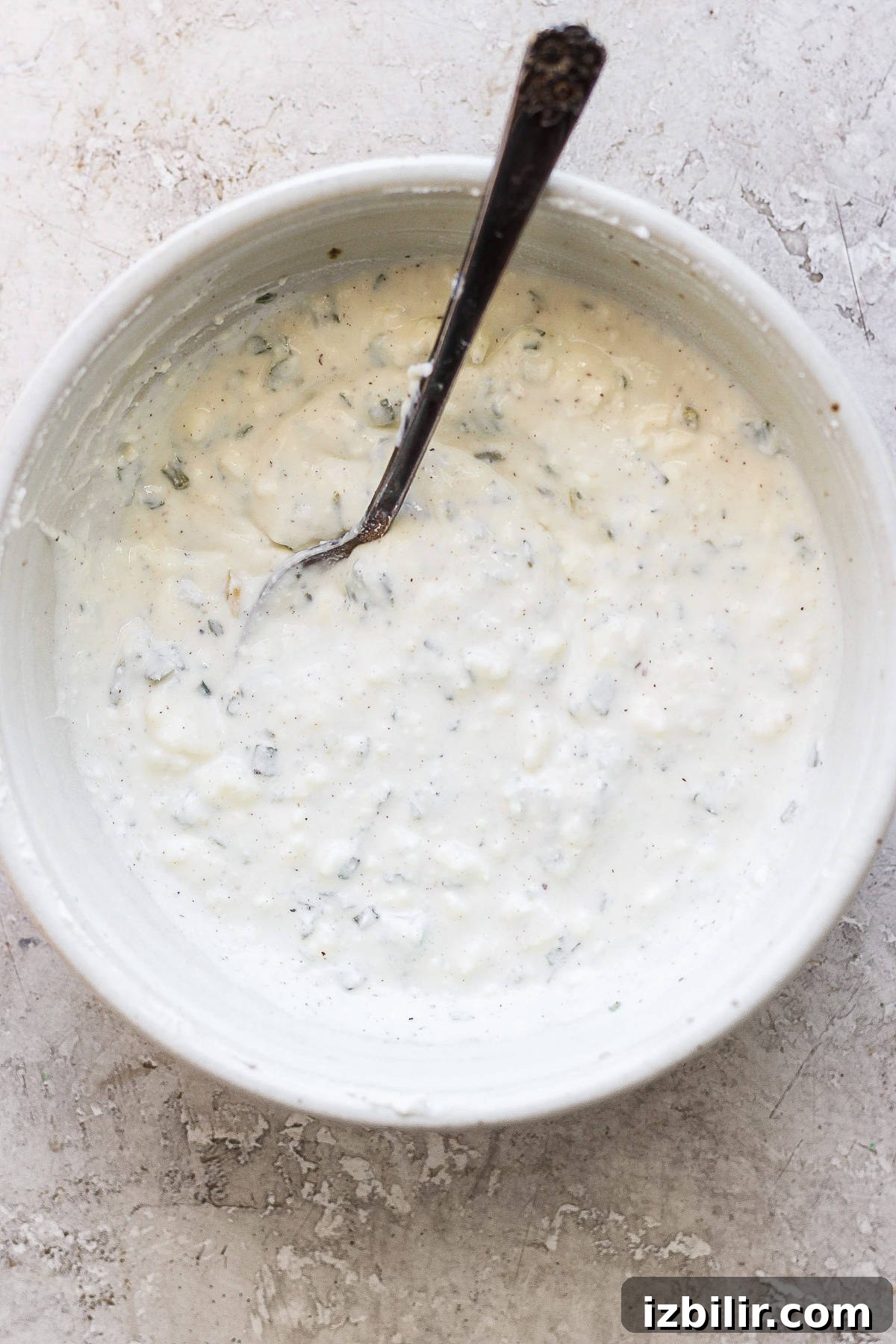 A bowl of creamy herb dressing with a spoon on a textured surface.