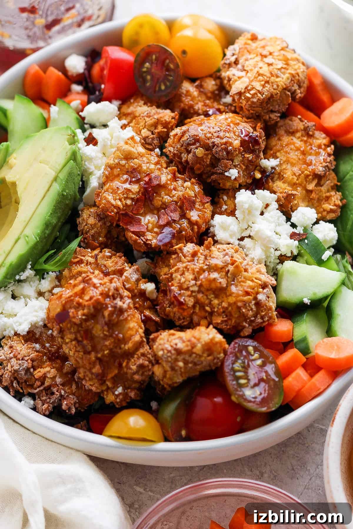 A bowl filled with crispy fried chicken, avocado slices, cherry tomatoes, cucumbers, carrots, and crumbled cheese, presenting a colorful and appetizing mixed meal.