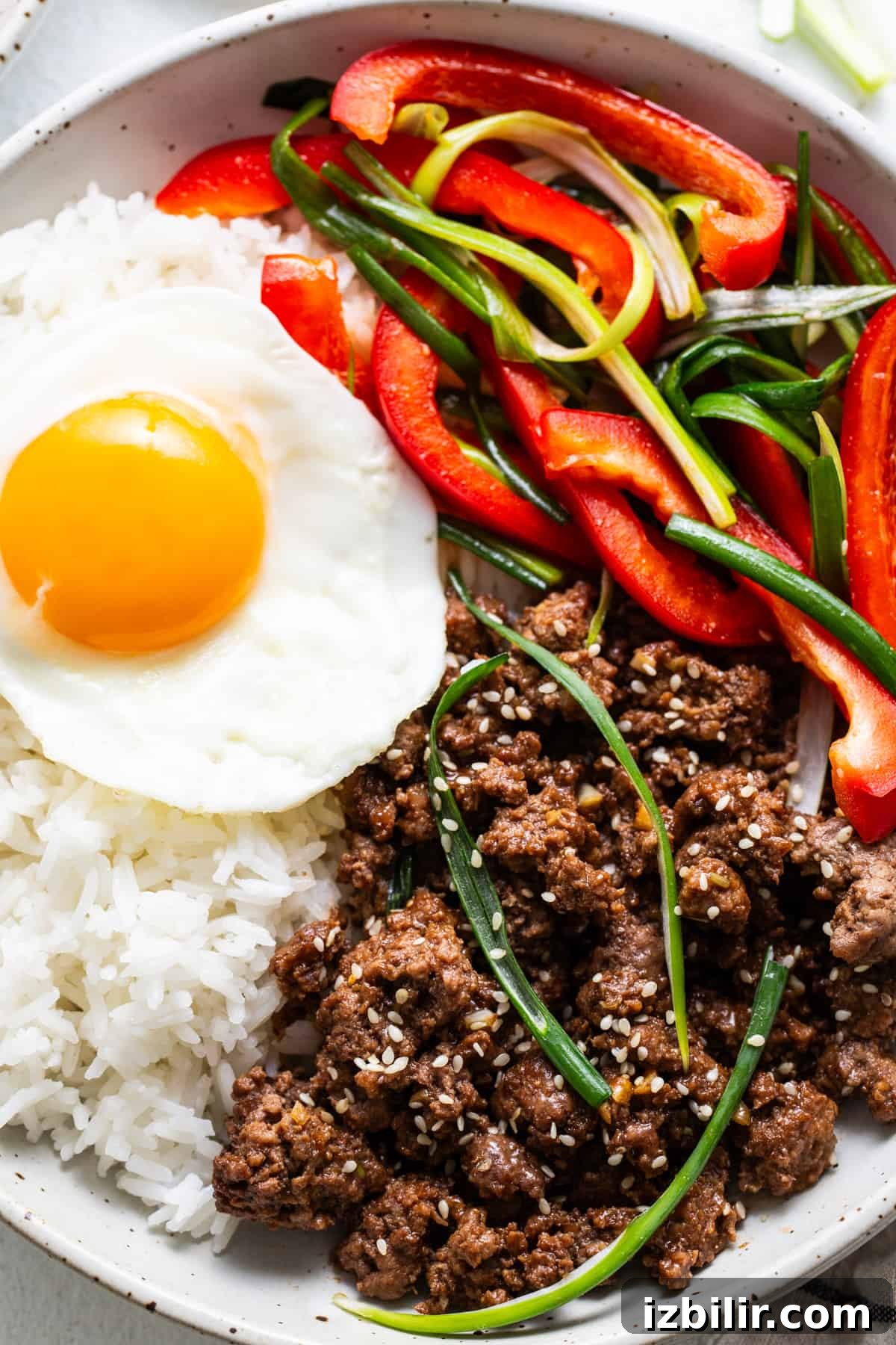 A bowl of white rice topped with a sunny side up egg, seasoned Korean ground beef, sliced red bell peppers, and green onions, sprinkled with sesame seeds.