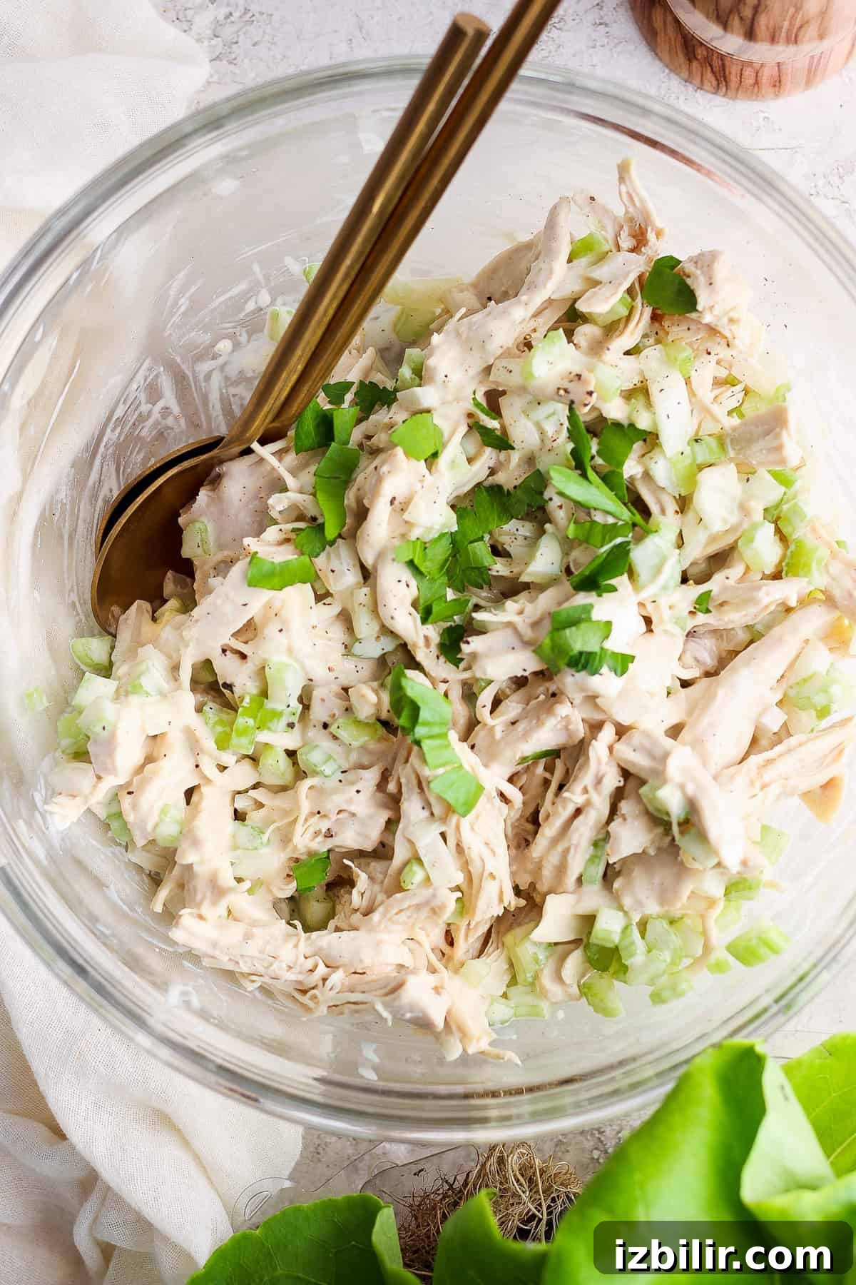 A bowl containing shredded chicken mixed with chopped celery and herbs, with a pair of gold-colored utensils.