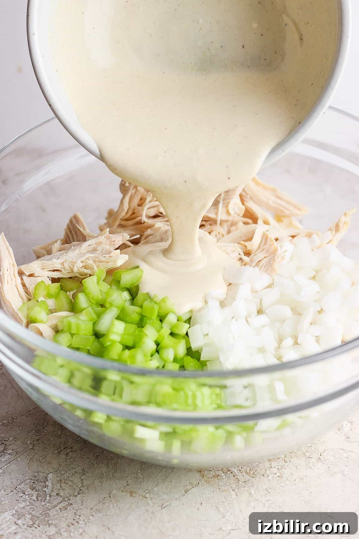 A creamy sauce is being poured into a bowl containing shredded chicken, diced onions, and chopped celery.