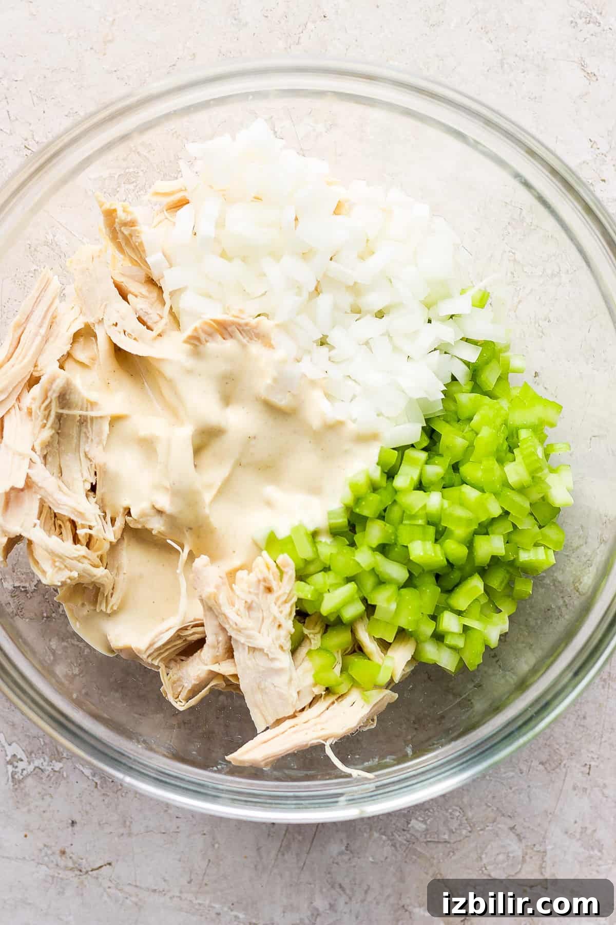 A glass bowl containing shredded chicken, diced onions, and chopped celery, possibly in preparation for a chicken salad or similar dish.