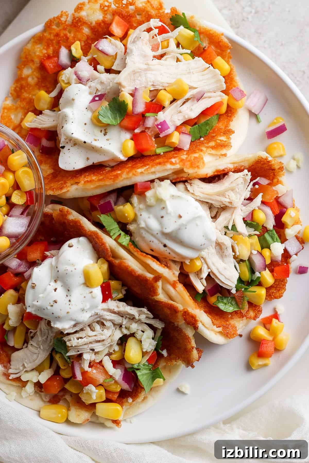Crispy Chicken Tacos with Cotija Cheese and Lime 2 A plate containing chicken tacos garnished with corn, diced tomatoes, red onion, cilantro, and a dollop of sour cream, served on a white surface.