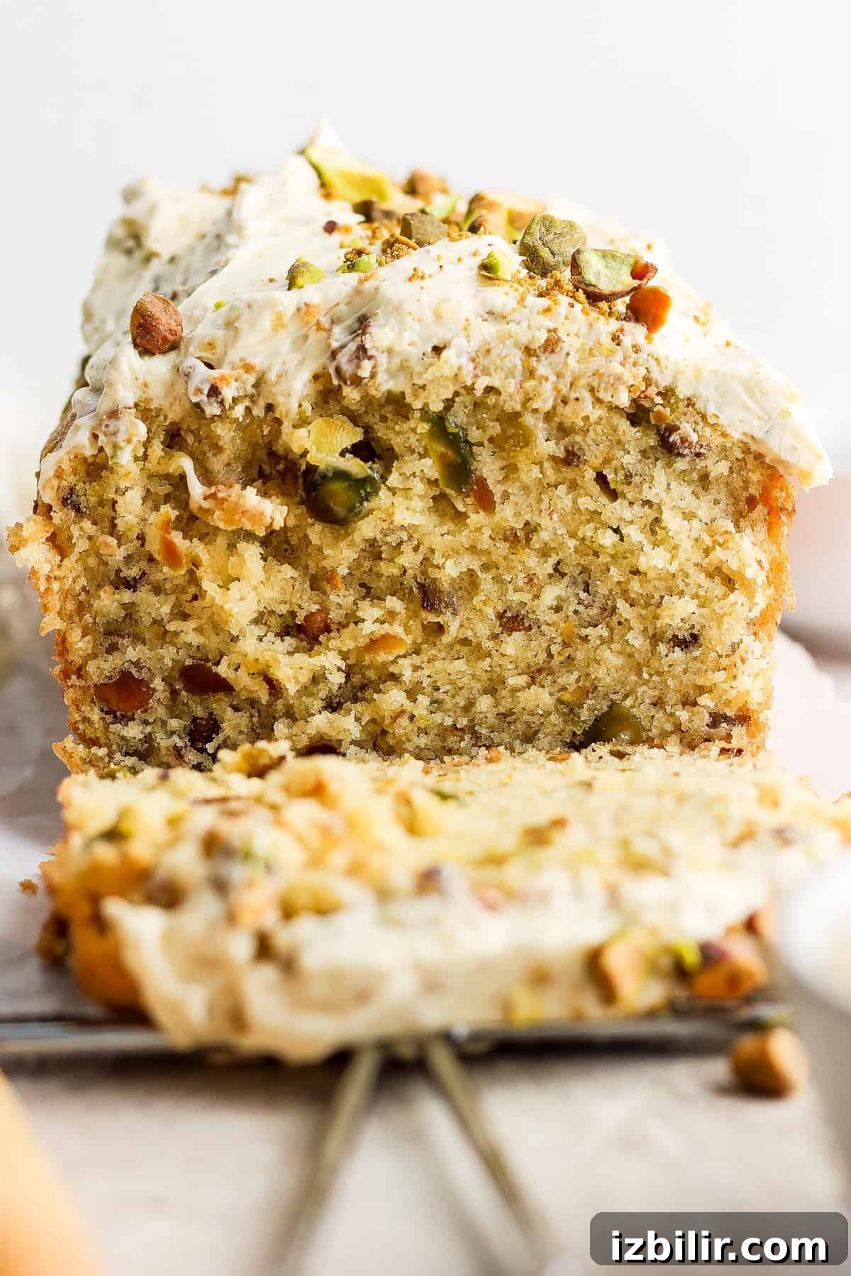 Vanilla Pistachio Loaf Recipe with Pistachio Frosting 2 A close-up of a slice of pistachio cake with frosting and chopped nuts on top, with the remaining cake in the background.