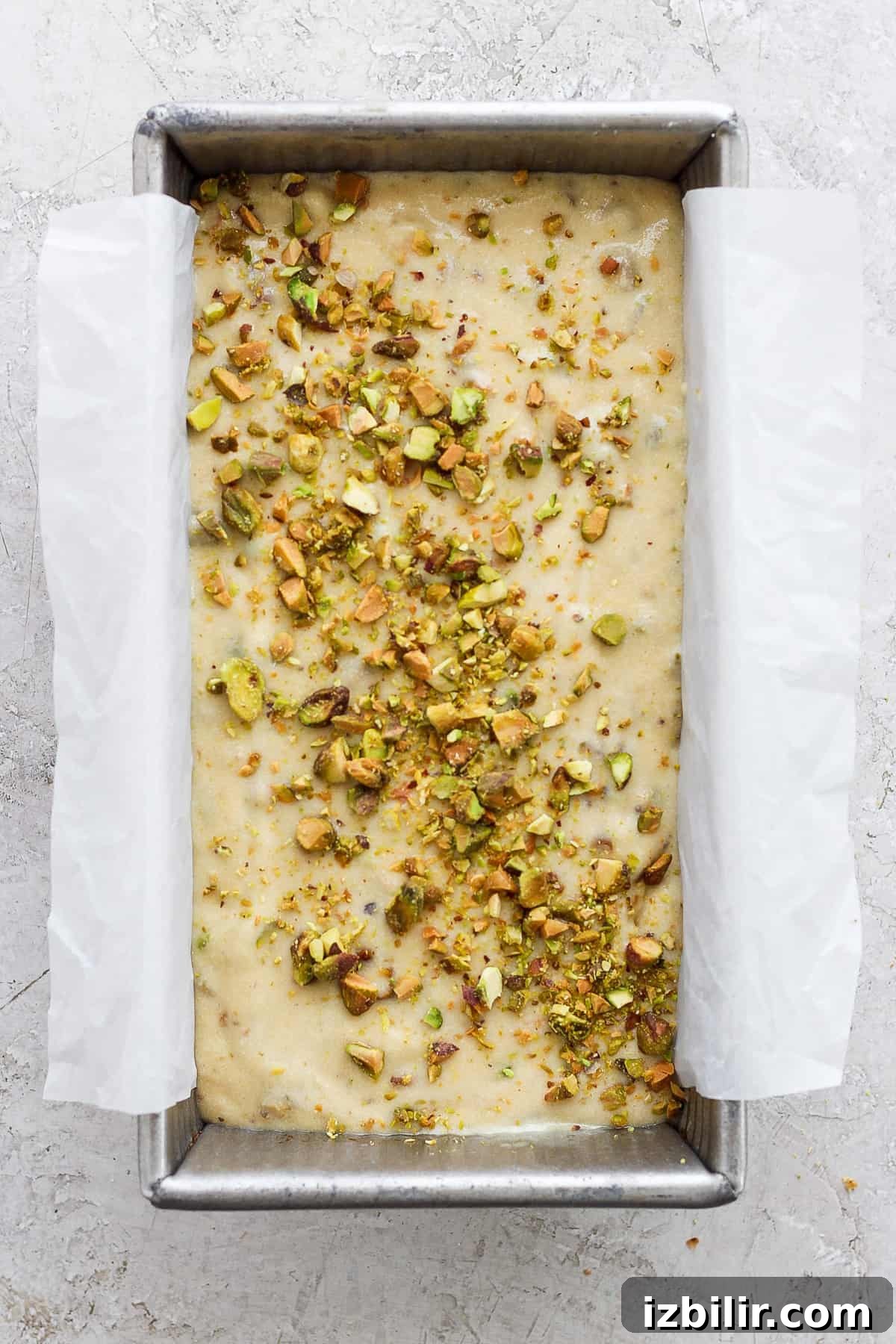 Vanilla Pistachio Loaf Recipe with Pistachio Frosting 3 Unbaked dough in a metal tray topped with chopped nuts, prepared for baking and lined with parchment paper.