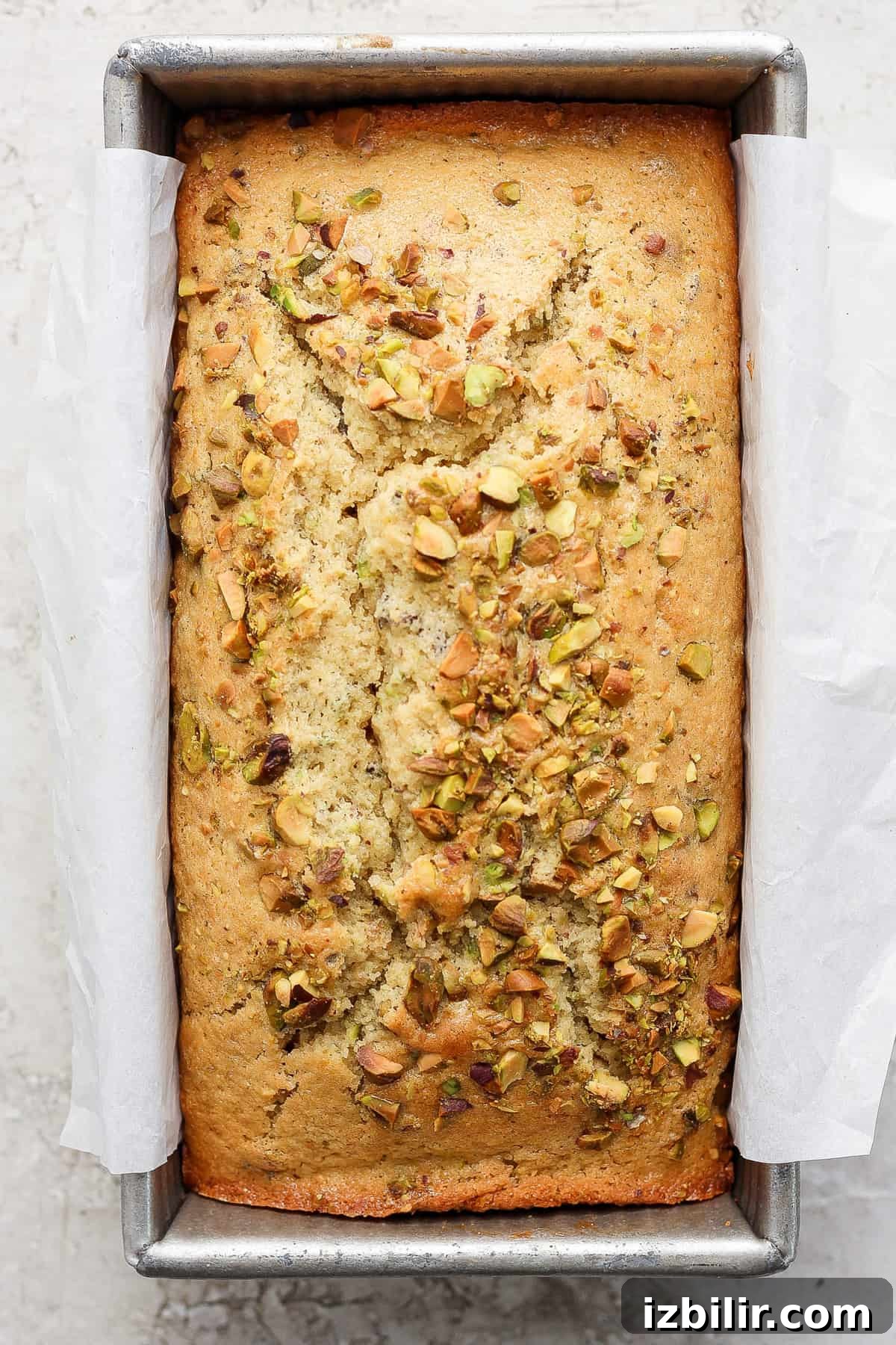 Vanilla Pistachio Loaf Recipe with Pistachio Frosting 4 A freshly-baked loaf of bread with pistachios sprinkled on top, sitting in a metal baking pan lined with parchment paper.
