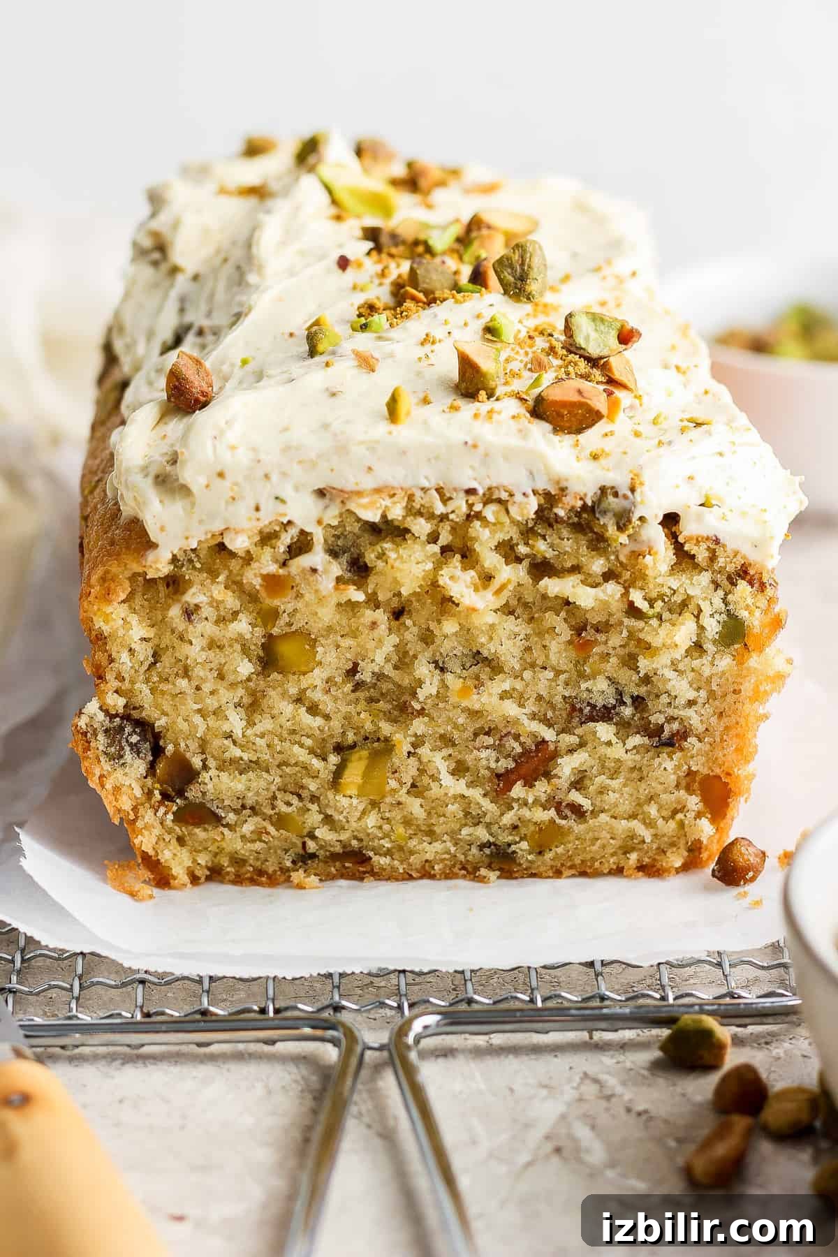 Vanilla Pistachio Loaf Recipe with Pistachio Frosting 6 A loaf of frosted cake with nuts and chopped pistachios on top, partially sliced, displayed on a piece of parchment paper.