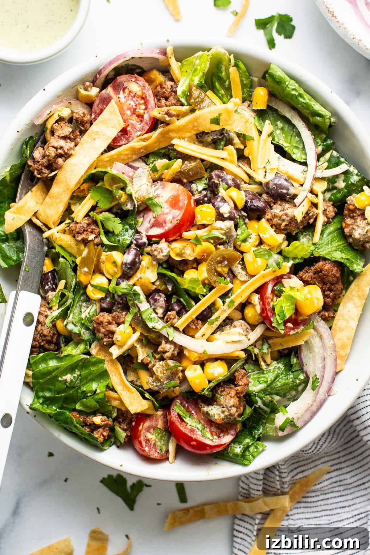 A vibrant taco salad with ground meat, mixed greens, tomatoes, cheese, corn, black beans, and crispy tortilla strips, served with a creamy dressing on the side.