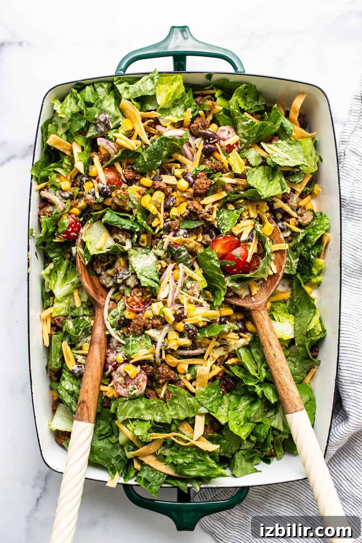 A colorful mixed salad with various greens, nuts, cheese, and tomatoes served in a large green dish with wooden serving utensils.