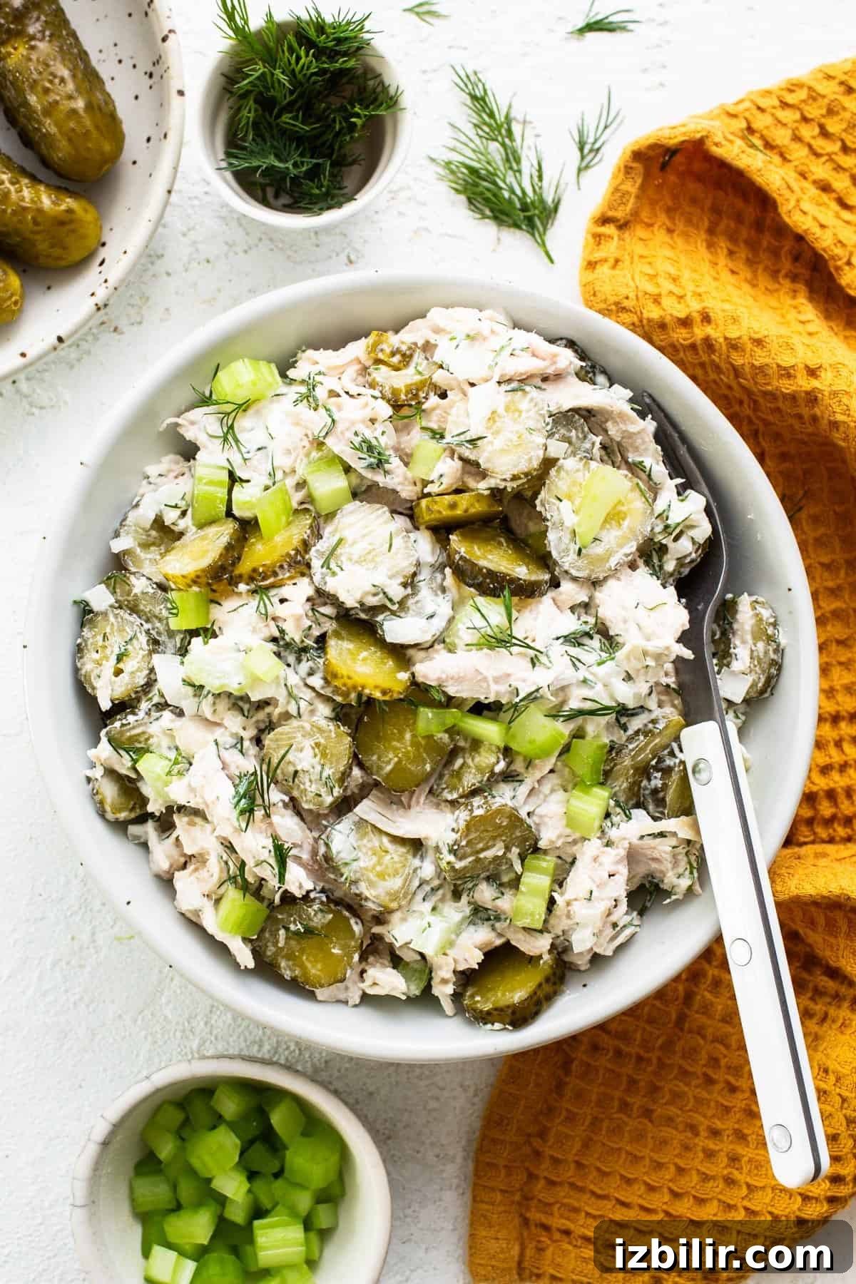 Fit Foodie Cooking Club April 2024 Classes and Recipes 6 A bowl of chicken and pickle salad garnished with dill, accompanied by a serving fork, on a white surface with pickles and chopped celery nearby.