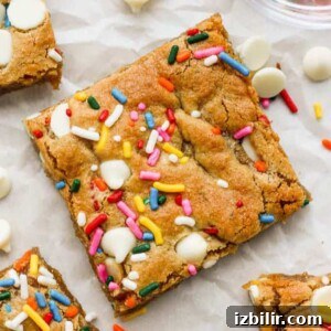 White Chocolate Funfetti Blondies Recipe 7 A close-up of a square Funfetti blondie topped with colorful sprinkles and white chocolate chips, with more scattered around it.
