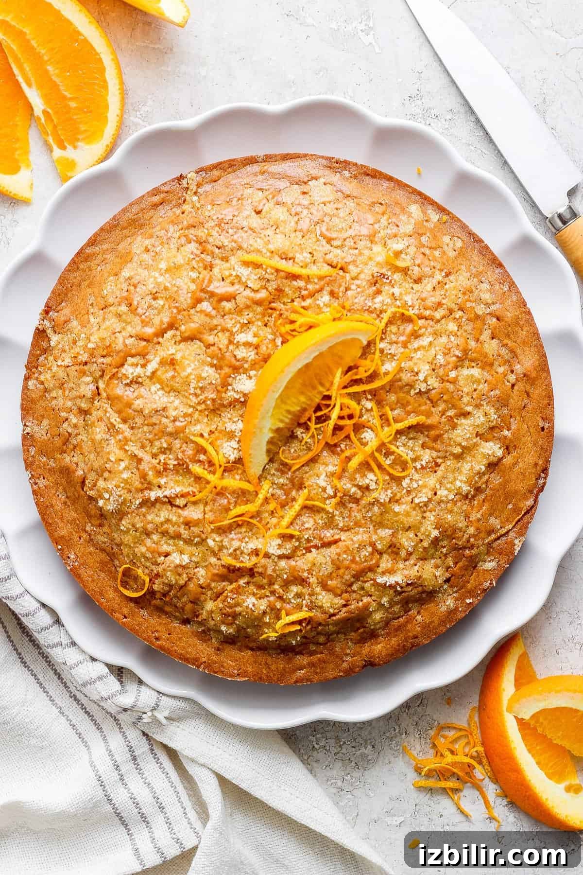 A freshly-baked orange cake garnished with orange zest on a white plate, accompanied by sliced oranges.